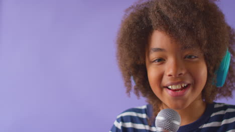 Foto-De-Estudio-De-Un-Niño-Con-Audífonos-Cantando-Karaoke-En-El-Micrófono-Contra-Un-Fondo-Morado