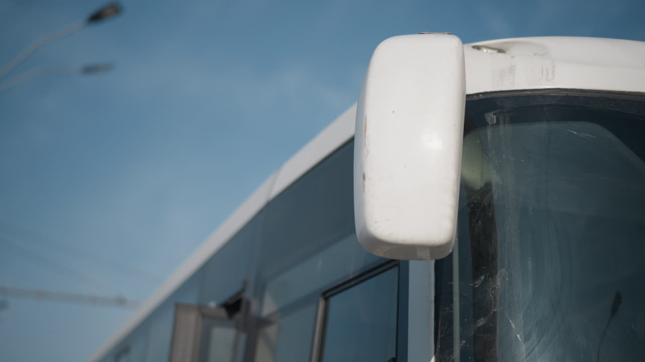 upward view shows modern passenger bus parked at stop, side mirror and curved front under blue sky, overhead power wires and street lamps in frame, calm urban transit moment, door closed, engine idle