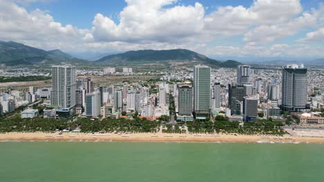 drone aéreo volando paralelo a la costa de la playa de arena blanca llena de palmeras y sombrillas de playa blanca en nha trang vietam en un día soleado con altos edificios residenciales a lo largo de una carretera costera