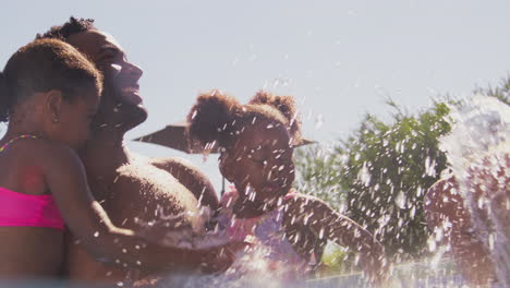 Family-On-Summer-Holiday-With-Two-Girls-In-Swimming-Pool-Splashing-Mother