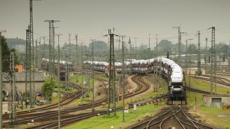 Handheld-camera-captures-a-train-yard-with-a-some-freight-trains,-encircled-by-trees,-buildings,-power-lines-under-an-overcast,-hazy-sky