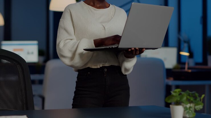 Afrikanischer Freiberufler lächelt in die Kamera, hält einen Laptop und steht neben einem Schreibtisch.