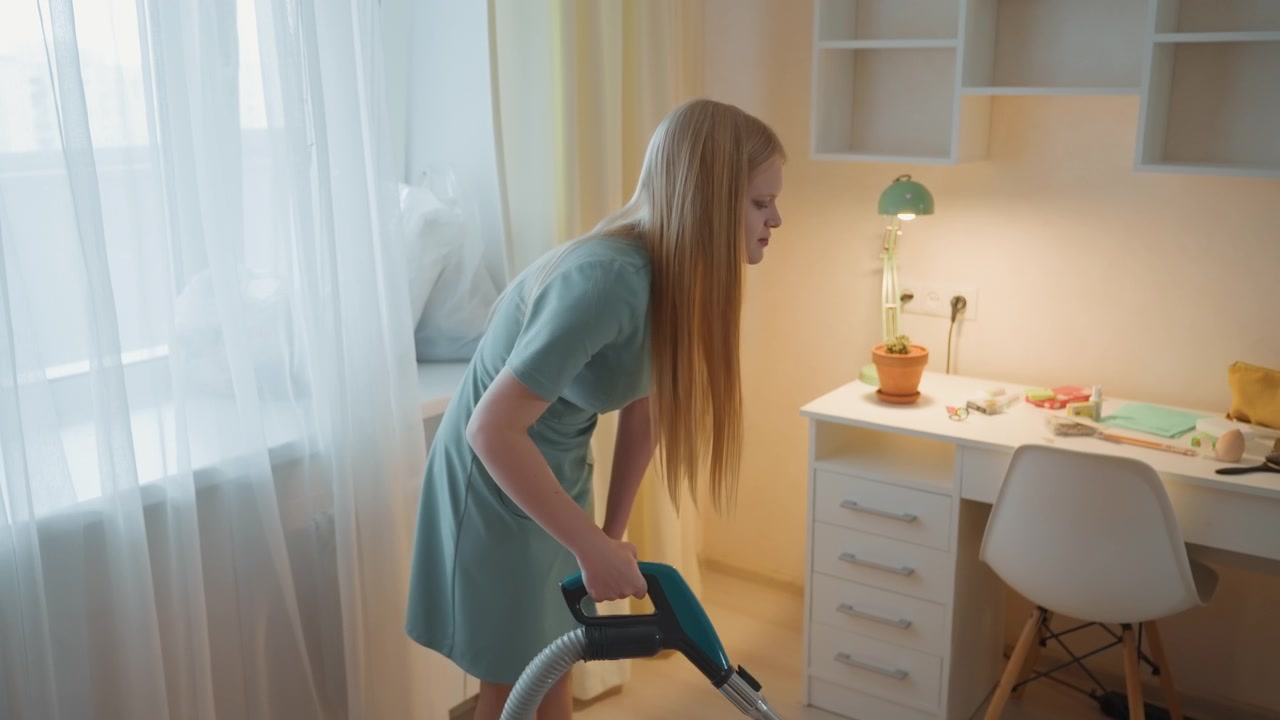 House cleaning young woman vacuuming desk in sunlit bedroom, focused tidy routine with white desk, lamp, chair and sheer curtains, small plant on desk, methodical motion and warm interior light