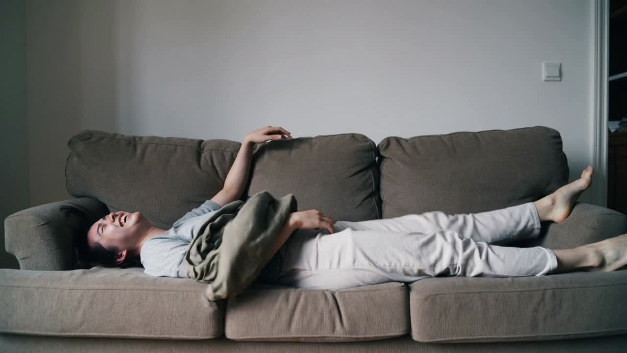 Spostando il peso, un uomo adulto in maglietta grigia si lascia cadere sul divano in salotto, prendendo una coperta per rilassarsi