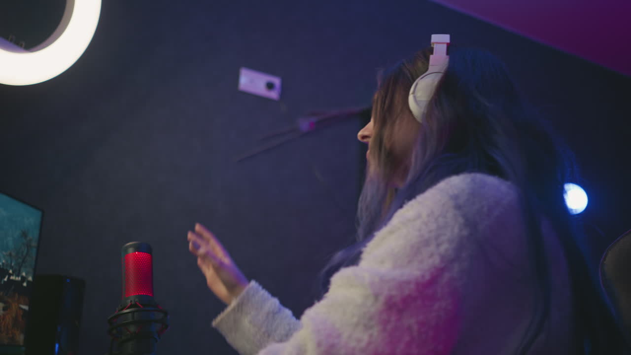 Top view of woman wearing white headphones concentrating on monitor in neon lit studio, expression shows disappointment while typing at keyboard beside red microphone on dark desk