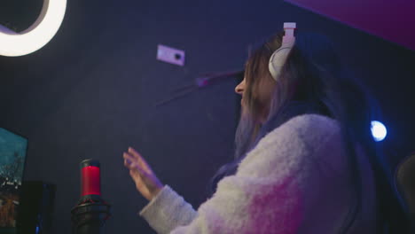 top view of woman wearing white headphones concentrating on monitor in neon lit studio, expression shows disappointment while typing at keyboard beside red microphone on dark desk