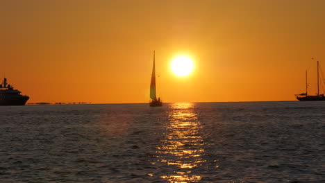 the sun sets behind a silhouetted sailboat with it's sail up of the coast of key west florida.