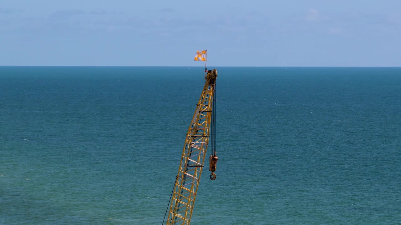 Side view of a tall metal construction crane boom with a hanging hook and checkered flag above a calm blue ocean.