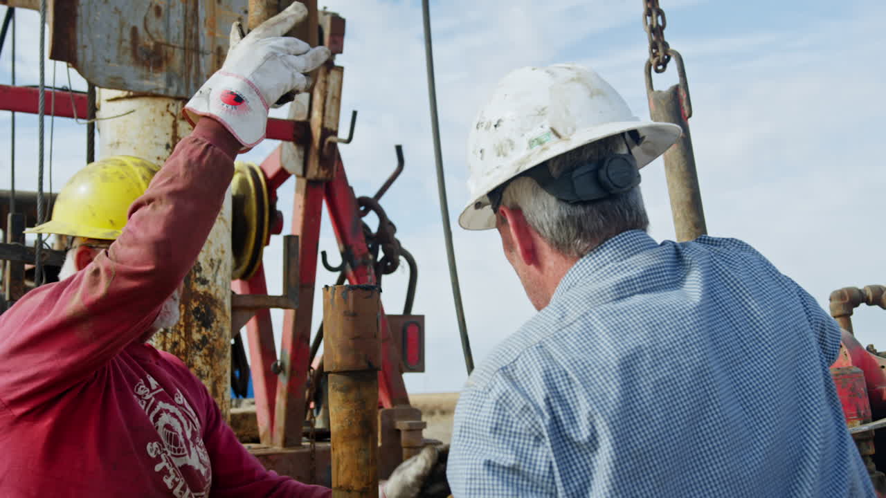 Hard-working men wearing helmets arrange equipment at oil production site. Workers fix metal pipes for drilling natural resources.