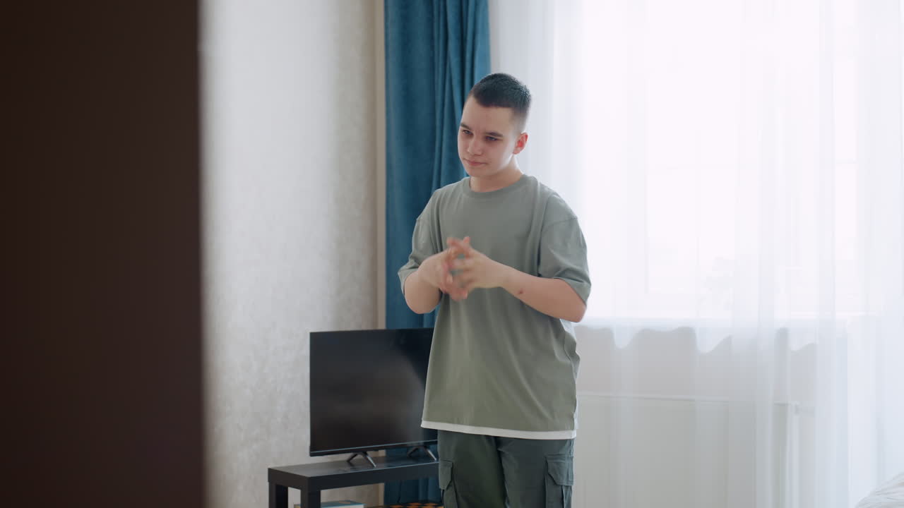 Confident young boy in casual outfit stands in bright room performing wrist twist exercise for hand flexibility and warm up, natural light from window and calm indoor setting