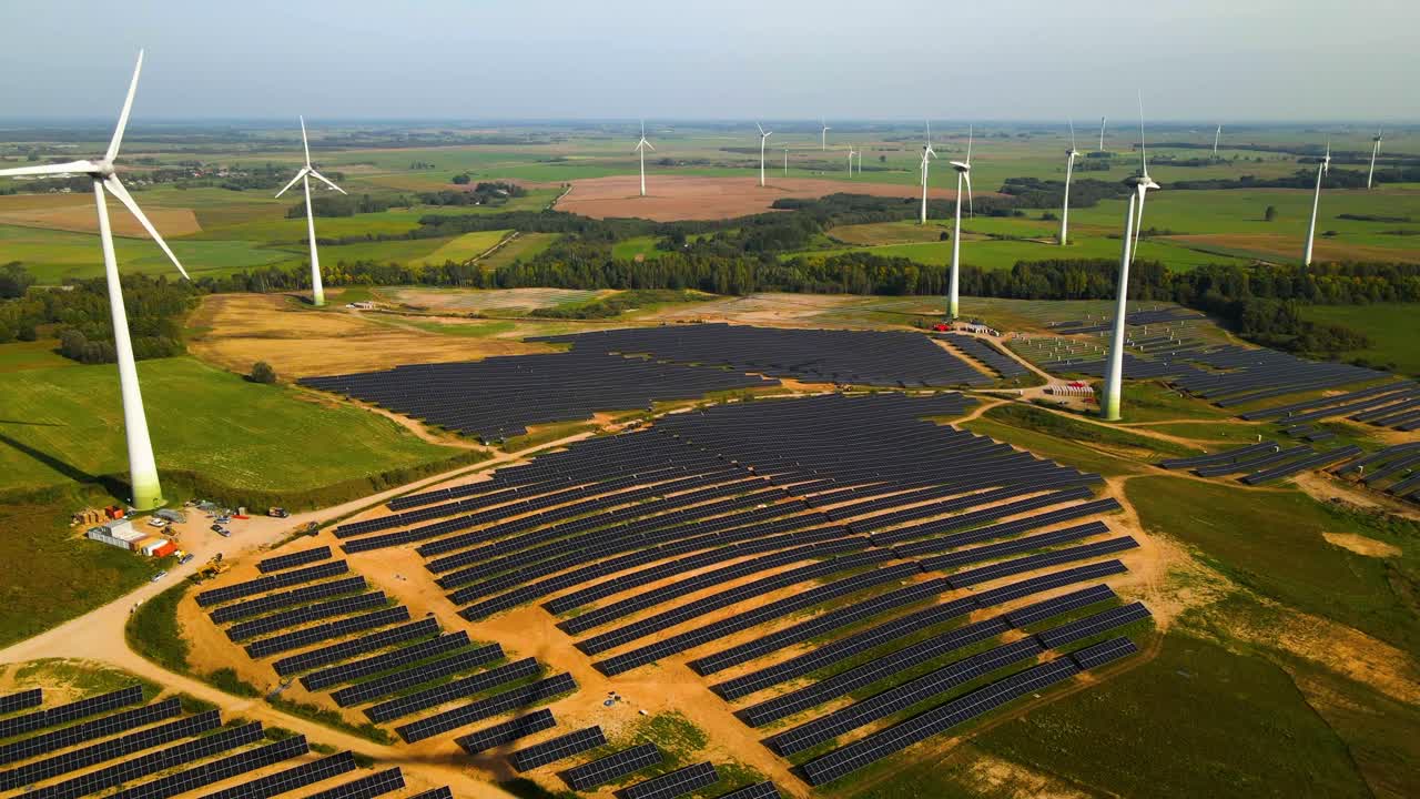 Aerial footage of solar panels plant and wind turbines in a wind farm generating green electric energy on a wide green field on a sunny day, in Taurage, Lithuania, zooming out