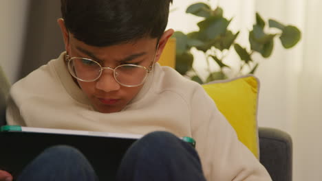 close up of young boy sitting on sofa at home playing games or streaming onto digital tablet