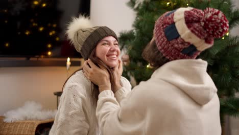 Happy-couple-a-guy-in-a-red-woolen-hat-and-a-brunette-girl-in-a-green-woolen-hat-and-a-white-sweater-adjust-their-hats-and-look-into-each-other's-eyes-in-a-cozy-room-decorated-with-the-universe-of-the-year-with-a-green-New-Year-tree