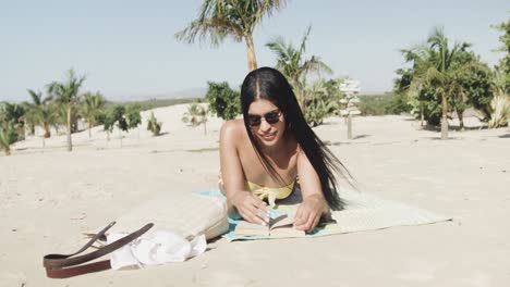 Fröhliche-Hispanische-Frau-Mit-Sonnenbrille,-Die-Am-Strand-Liegt-Und-Ein-Buch-In-Der-Sonne-Liest,-Zeitlupe