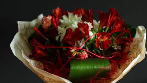Ein-Großer-Strauß-Aus-Roten-Und-Weißen-Blumen-Und-Grünen-Zierelementen-Dreht-Sich-In-Einer-Mit-Papier-Dekorierten-Glasvase-Auf-Dunklem-Hintergrund
