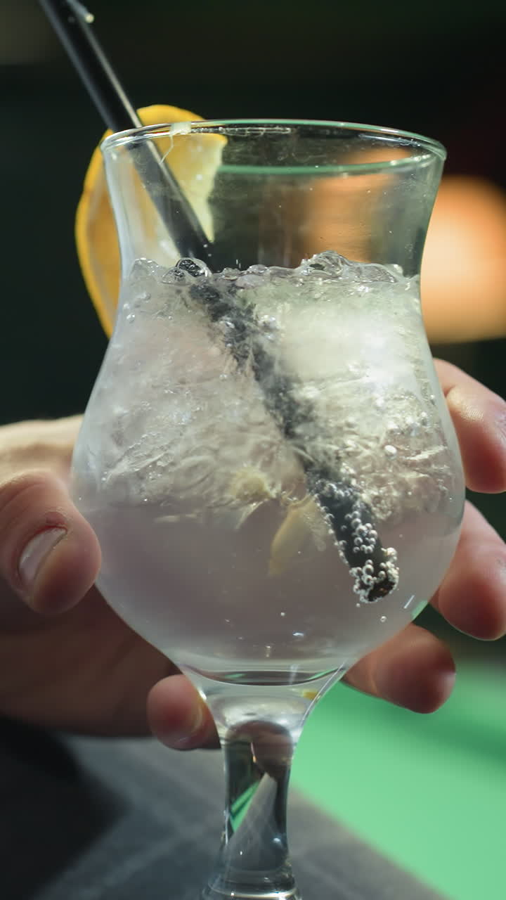 primer plano de una persona, cara no visible, con camisa blanca y pantalones grises, sosteniendo un vaso de bebida de limón con paja negra. cubos de hielo y rebanada de limón