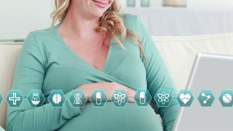 Medical-icons-over-happy-pregnant-caucasian-woman-on-couch-using-laptop