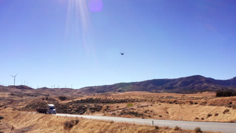 un camión conduce a lo largo de la carretera del desierto con turbinas eólicas a lo largo de las colinas en el fondo - vista aérea deslizante