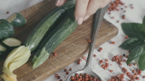 bodegón vegetal: una mano coge un calabacín que descansa sobre una tabla de madera, en la composición de la escena hay pimiento rojo en una cuchara de plata y hojas de albahaca