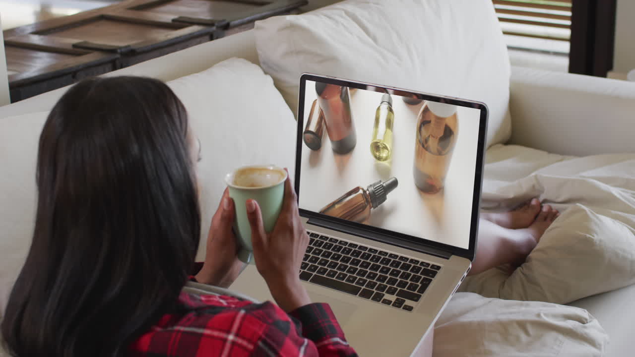 Biracial woman using laptop on couch at home online shopping for beauty products, slow motion