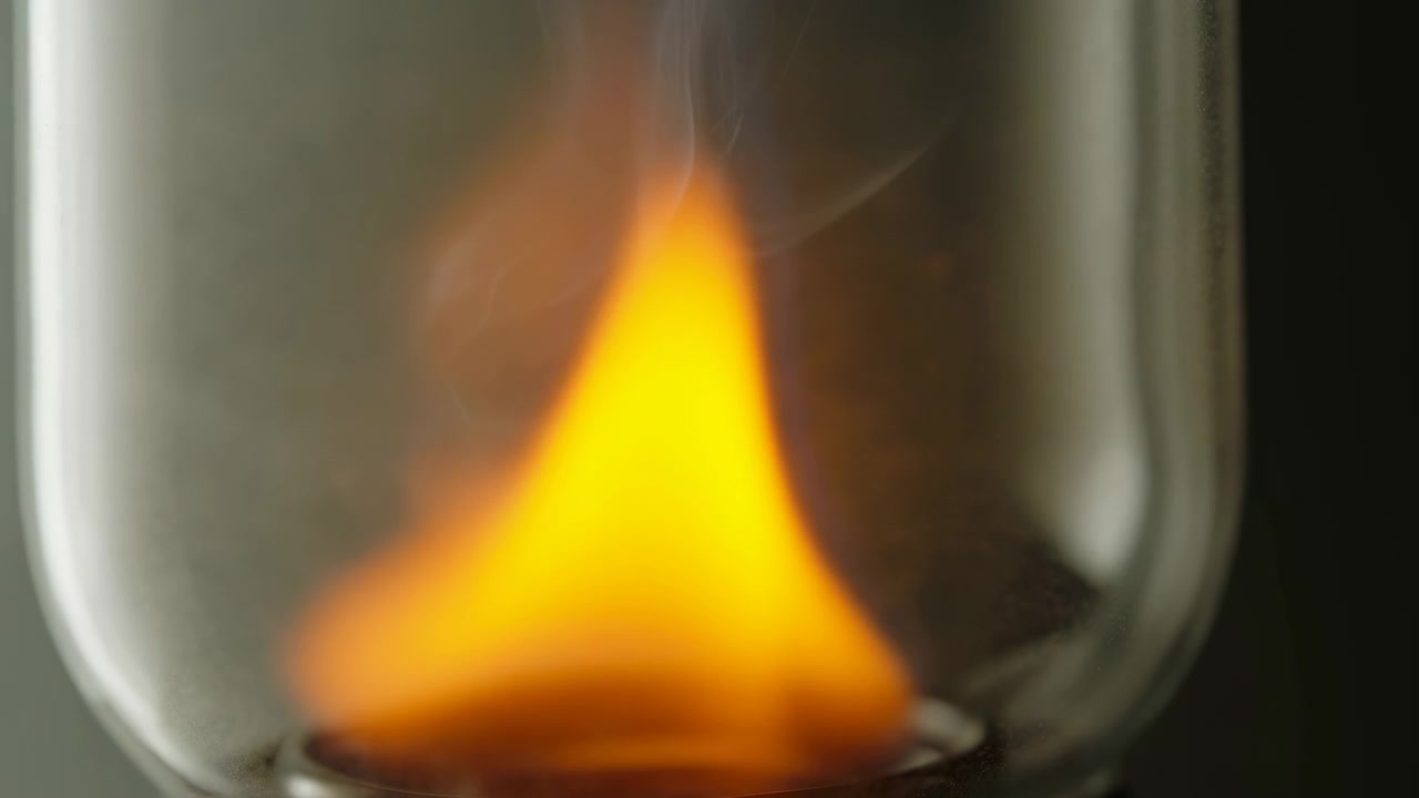Un primo piano intimo mostra una fiamma giallo-arancio brillante che brucia all'interno di un vetro trasparente curvo, che si innalza con una punta affusolata mentre deboli volute di fumo si librano sopra, con una base dello stoppino scura e tenui riflessi intorno ad essa.