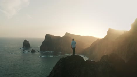 young male adventure hiker reaching top of wild viewpoint, looking at rugged coastal cliffs with bright morning sunlight, successful summit, aerial