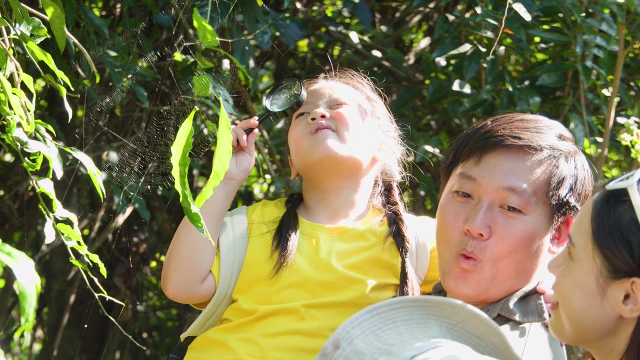 タイ人家族が緑豊かな森を探索し、幼い娘が虫眼鏡で緑の葉を観察している。