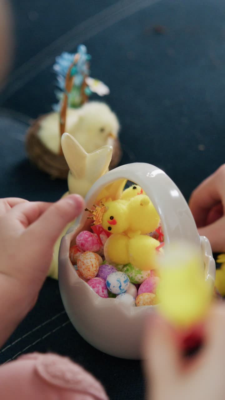 Hands gently place tiny yellow chicks into a ceramic egg filled with colorful candies. The scene captures joyful, playful interaction during Easter preparations. Soft focus highlights the vibrant