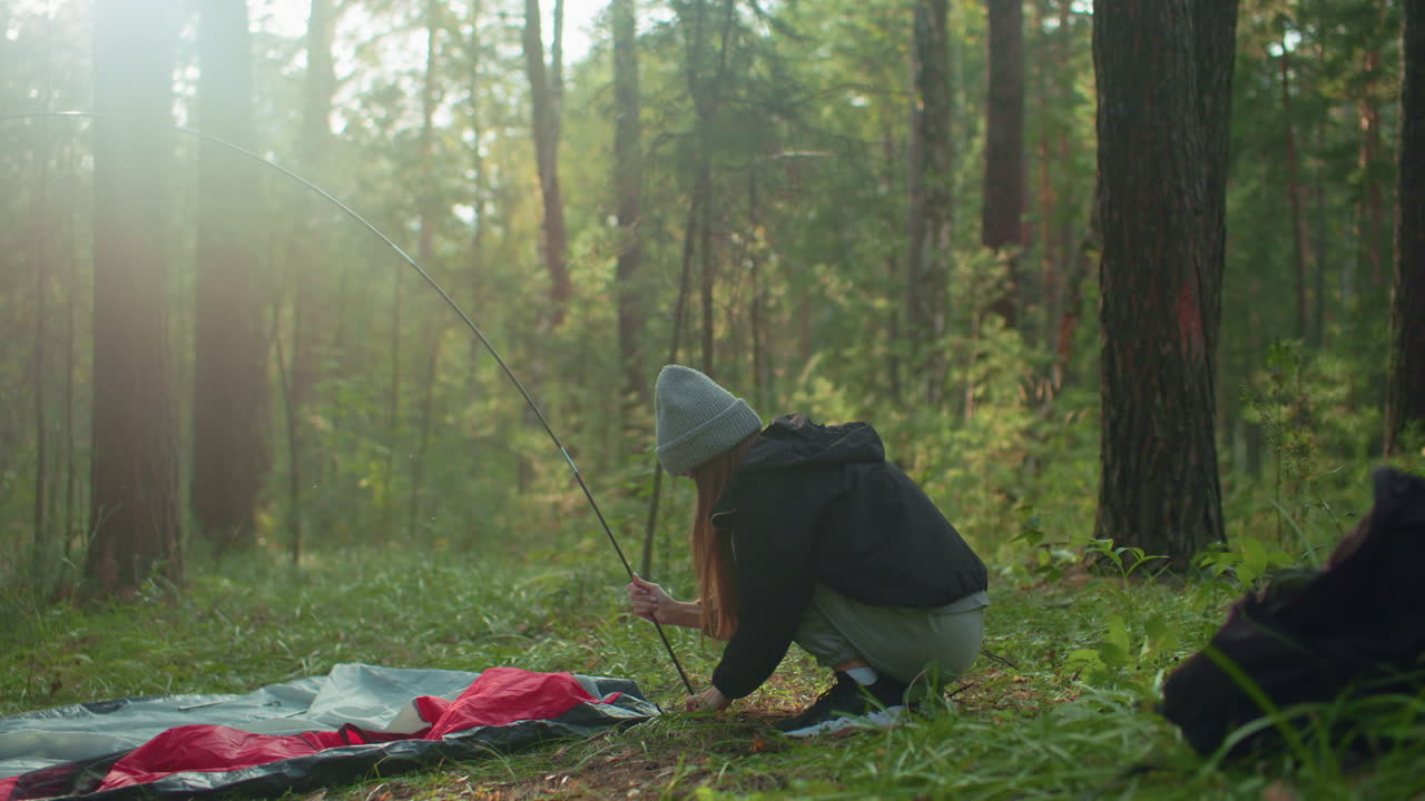 Woman squats in forest holding flexible tent pole while lifting tent fabric to secure around base, sunlight filters through trees casting gentle glow over peaceful camping moment surrounded by trees