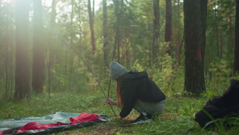 woman squats in forest holding flexible tent pole while lifting tent fabric to secure around base, sunlight filters through trees casting gentle glow over peaceful camping moment surrounded by trees