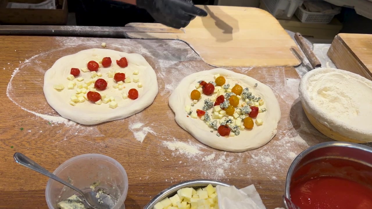 Ripresa dall'alto di uno chef con guanti neri che aggiunge formaggio e pomodori all'impasto della pizza
