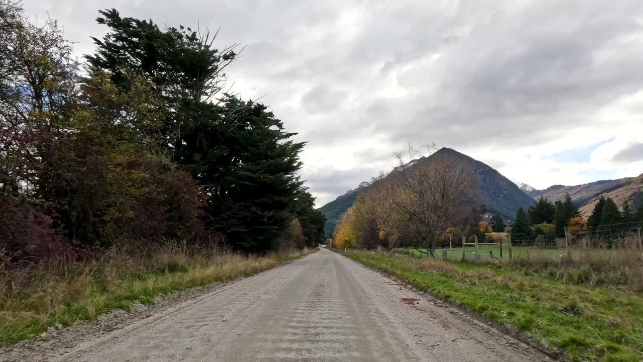 Vehicle travels rural gravel road, passing trees and fields under overcast sky, steady forward motion