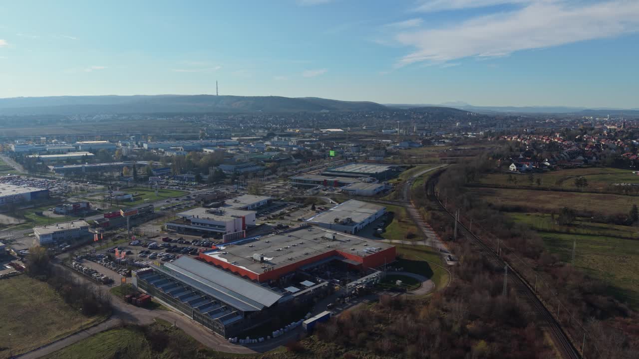 A wide aerial view showing an industrial and commercial district with large retail buildings, parking areas, and nearby roads. The scene extends toward distant hills and residential neighborhoods