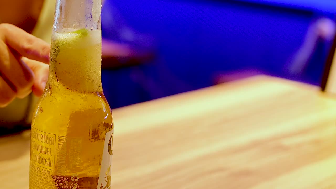 Close-up of a hand grasping a beer bottle on a wooden table with blue seating in the background.