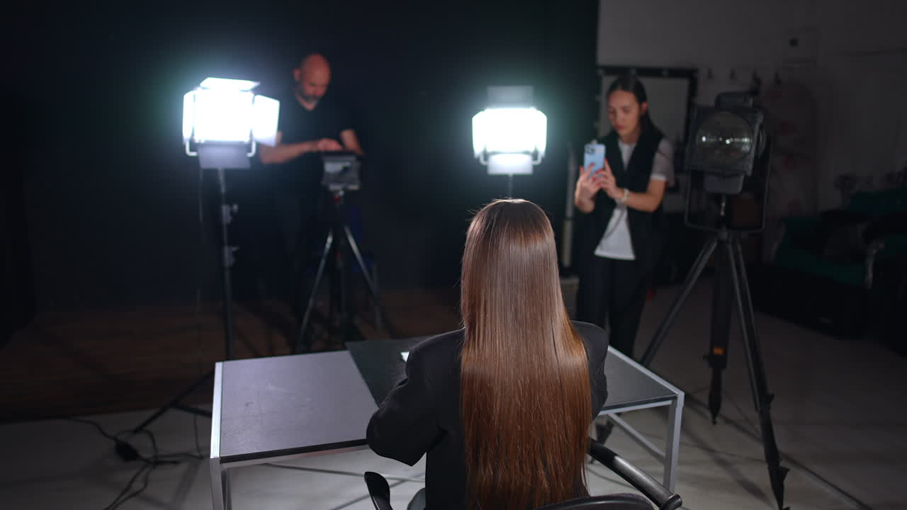 Rear view of a lady with long straight brunette hair sits at desk talking and gesturing in front of camera. Lights of soffits are on the reporter. Cameraman and girl filming on her phone at backdrop.