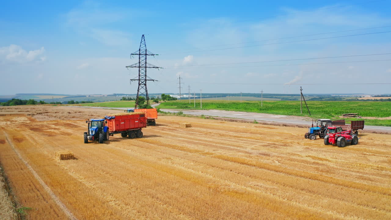 Loaded tractor full of straw leaves the field. Other agricultural machinery standing in the farmlands. Green plantations at backdrop.