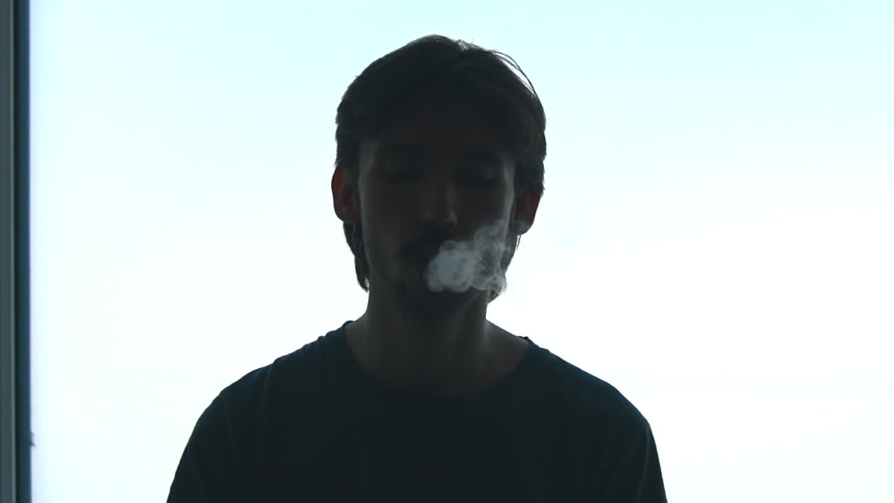 Uomo caucasico controluce dalla finestra che espira vapore durante la tranquilla routine mattutina, atmosfera da studente contemplativo. Stabile. Media.