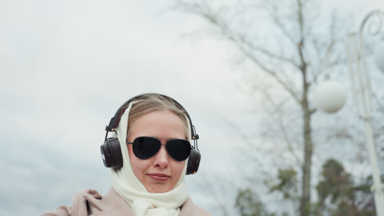 Donna nel parco che balla giocosamente a ritmo di musica, lanciando la borsa e ridendo, con cuffie e occhiali da sole. Alberi spogli e cielo nuvoloso fanno da sfondo. Un movimento energico e spontaneo e un'atmosfera da street fashion moderno.