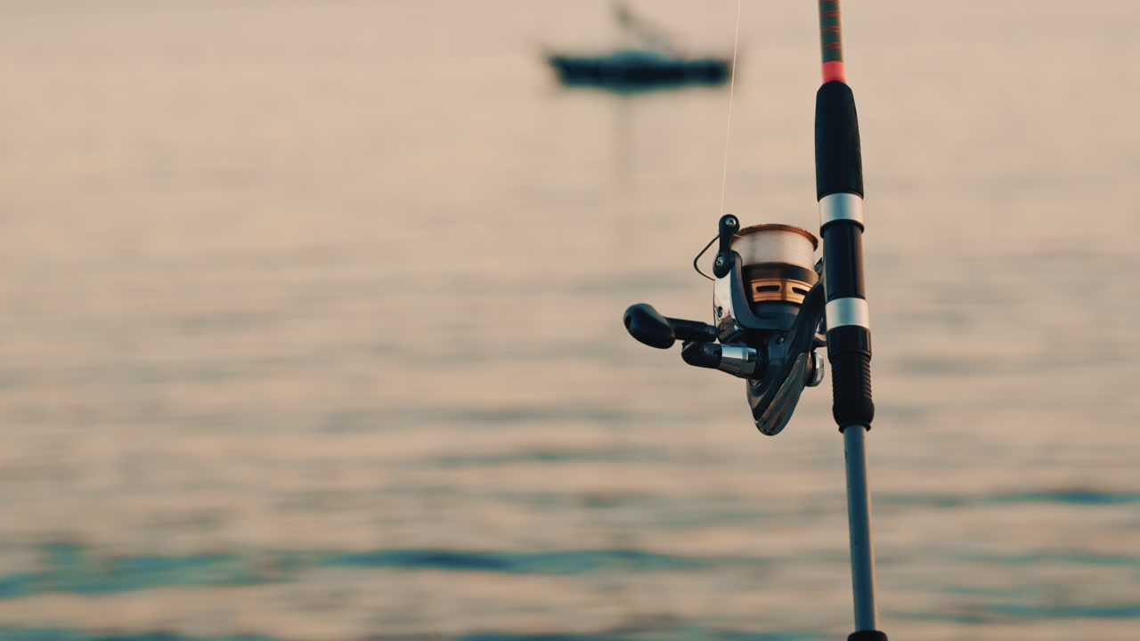Primo piano di una canna da pesca e un mulinello posizionati vicino al mare, con una barca sfocata sullo sfondo