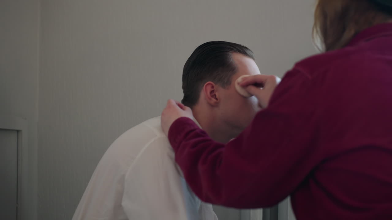 makeup artist applies products on man face during session as he closes eyes enjoying gentle cosmetic treatment in bright minimal studio setting with professional focus on skin blending technique