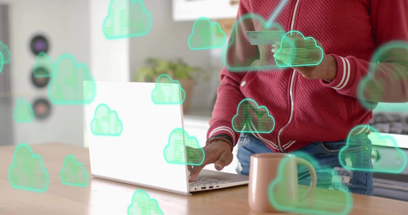 Donna che tiene una carta di pagamento e digita sul laptop in cucina, caricando dati di pagamento con icone cloud.