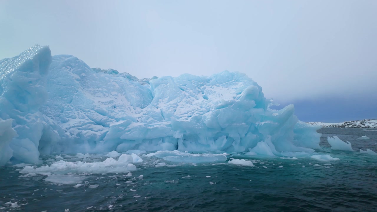 Blue iceberg slowly melting by Greenland's coast on calm, overcast day