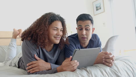 Happy-diverse-gay-male-couple-lying-on-bed-using-tablet-and-talking-in-the-morning,-slow-motion