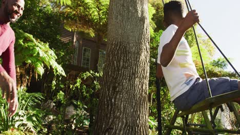 african american dad pushing his son on a swing in the garden on a bright sunny day