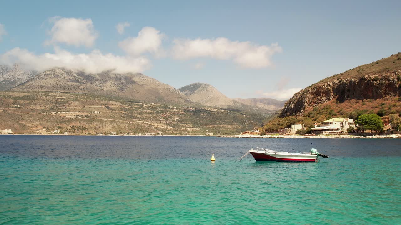 parallax aerial movement close to the sea level reveals small fishing boats while the camera gains altitude on a summer day with few clouds in the background covering the mountain side
