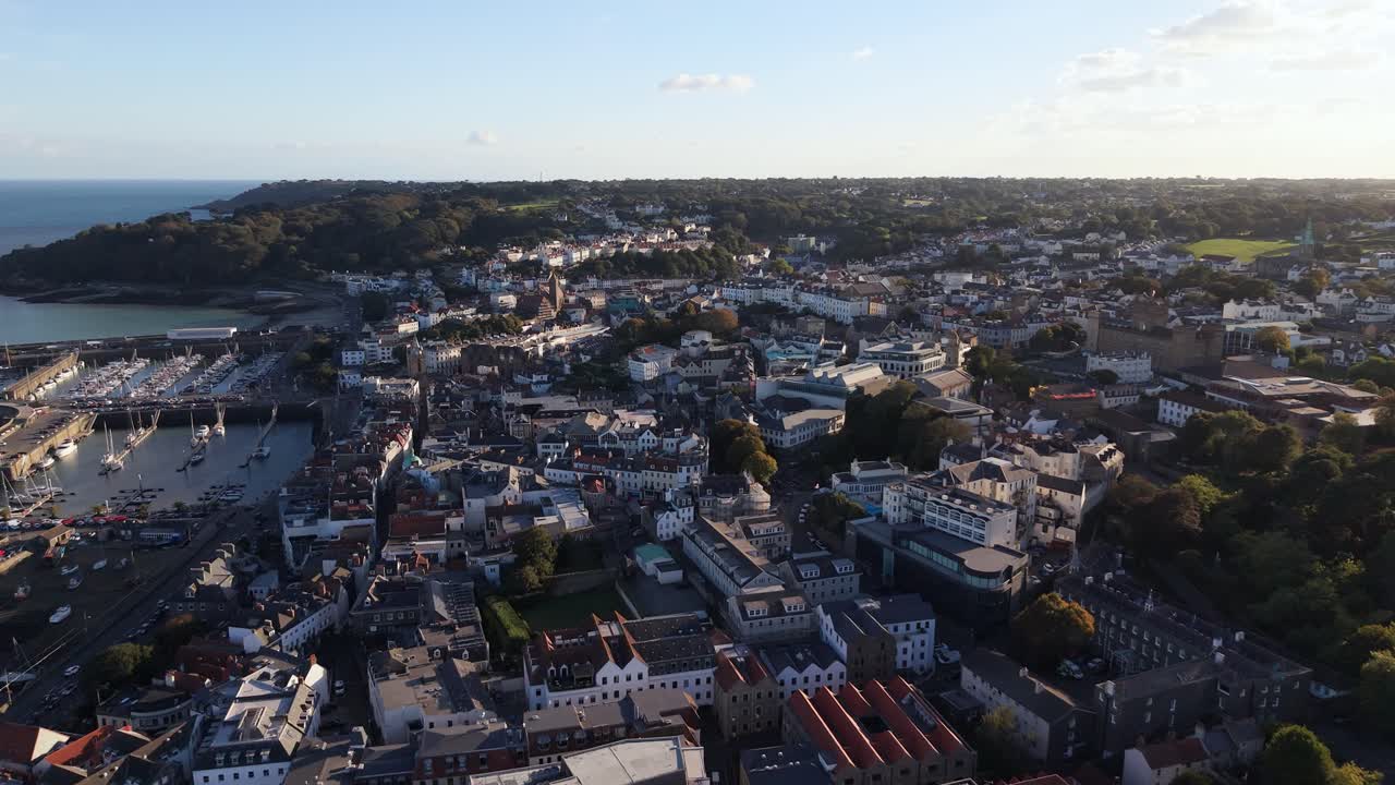 Slow drone footage of St Peter Port Guernsey including harbour and Havelet Bay high but detailed flying south with wooded cliffs and sea in the distance in late afternoon sun