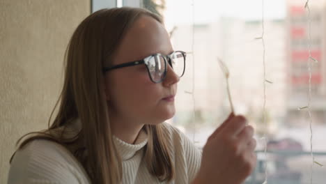 young woman in glasses joyfully licks spoon after savoring dessert by bright window with decorative lights and soft snowy background