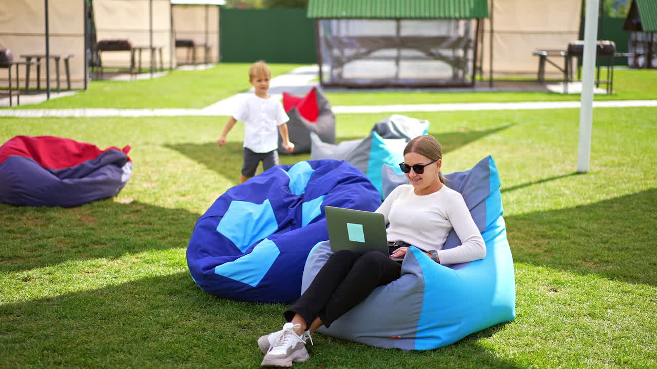 Blonde lady sitting in a bean bag chair works on laptop. Little boy comes up sitting down next, talking and hiding face in chair.