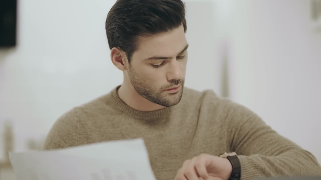 Tired white man working laptop at home kitchen. Surprised guy looking documents