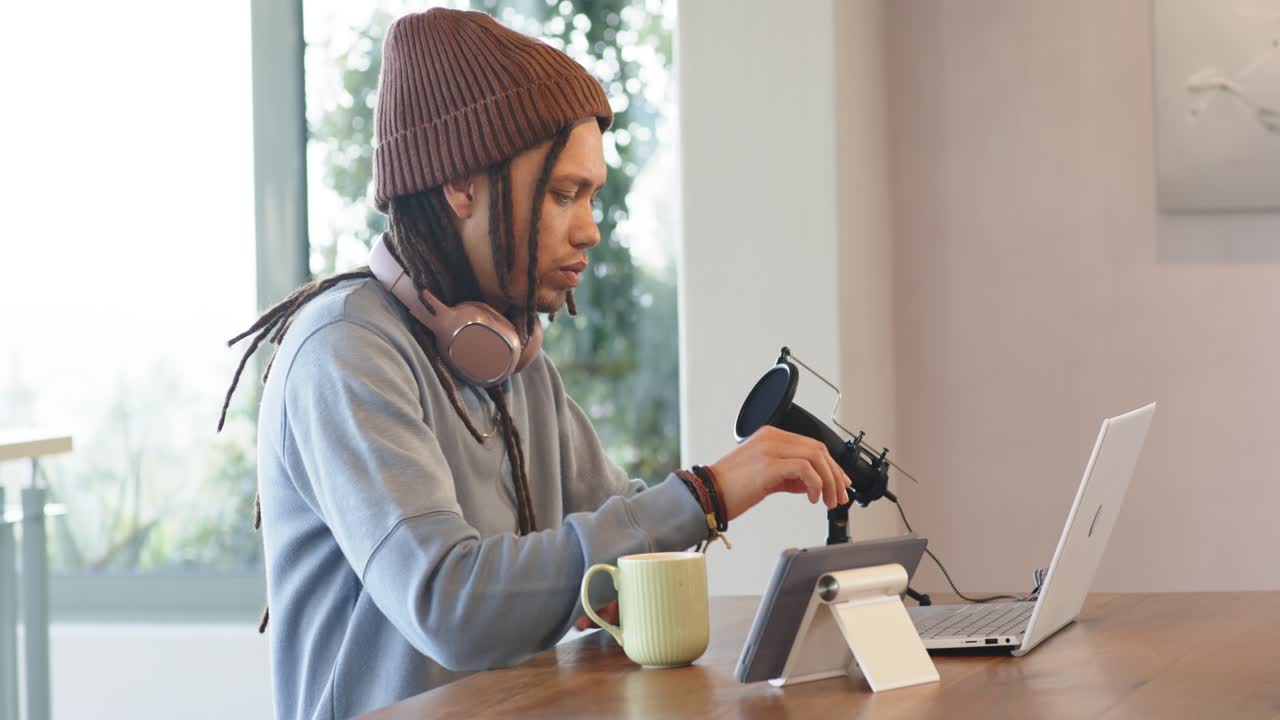Uomo seduto composto alla scrivania con un laptop in un ufficio domestico, che sorseggia una tazza, regola il microfono e scrive per lo streaming